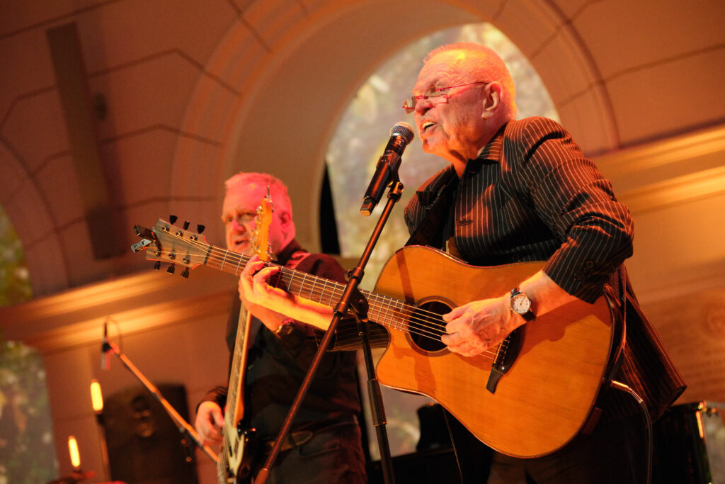 Jerzy Filar grający na gitarze i śpiewający podczas koncertu na Gali 75-lecia Ruchu Studenckiego w Auli Głównej Politechniki Warszawskiej, w towarzystwie basisty.